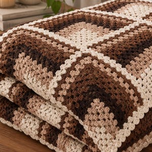 Può includere: Una coperta all'uncinetto piegata con un motivo geometrico a piastrelle granny nei toni del crema, marrone chiaro, marrone medio e marrone cioccolato scuro. Il tessuto è composto da motivi quadrati interconnessi con una trama di filato visibile.