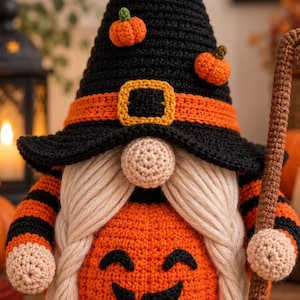 Puede incluir: Una figura de gnomo de ganchillo con un sombrero de bruja negro alto decorado con pequeñas calabazas naranjas y una banda naranja con una hebilla amarilla. El gnomo tiene una nariz beige, largas trenzas de hilo crema y un cuerpo de calabaza.