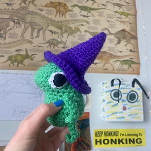 Könnte beinhalten: Eine handgefertigte grüne Häkelfrosch, der einen lila Hexenhut trägt. Der Frosch hat ein weißes und schwarzes Auge. Der Hintergrund umfasst ein Dinosaurierposter und ein gelbes Schild mit der Aufschrift "KEEP HONKING! I'm Listening To HONKING."