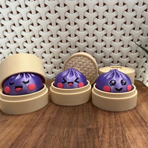 Pode incluir: Três figuras esféricas de bolinhos roxos impressas em 3D com expressões faciais distintas e bochechas cor-de-rosa, cada uma descansando dentro de um recipiente bege cilíndrico de duas peças que se assemelha a uma cesta de vapor.