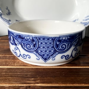 May include: A round ceramic bowl featuring a white base with a deep blue decorative pattern around the exterior. The design includes intricate floral motifs, scrolling lines, and a central geometric lattice panel. The bowl has a smooth rim.