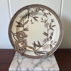 May include: A decorative ceramic plate with a bird and floral design in brown on a cream background. The plate has a detailed border and is displayed on a marble stand. The plate is approximately 25 cm in diameter.