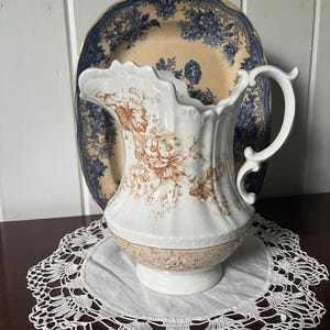 May include: A white ceramic pitcher featuring a brown floral transferware pattern with a scalloped rim and an ornate handle, positioned in front of a round plate with a blue floral pattern on a cream background, resting on a white lace doily.