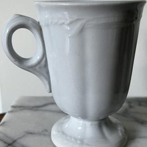 Puede incluir: Una taza de cerámica gris claro con base de pedestal y asa curva. La taza tiene un diseño acanalado en el cuerpo y un patrón de hojas decorativas cerca del borde. La taza probablemente se usa para bebidas.