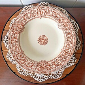 May include: A round ceramic soup bowl featuring a cream-colored base with a detailed reddish-brown transferware pattern. The design includes intricate scrollwork, floral motifs, and geometric borders along the rim, with a matching circular emblem in the center.