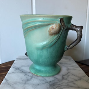 May include: A teal ceramic mug with a distinctive handle and decorative elements. The mug features a pedestal base and a textured finish. The handle is brown, and the design on the mug is a lighter shade of teal.