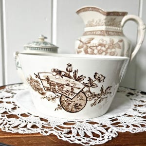 May include: A white ceramic bowl featuring a brown transferware pattern of a fan-shaped landscape illustration, floral motifs, and a circular medallion. The bowl has a smooth, tapered rim and a clean, rounded base, showcasing a detailed vintage aesthetic.