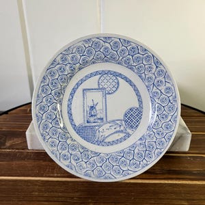 May include: A round, white and blue decorative plate with a windmill scene in the center. The plate has a blue patterned border and a blue and white design around the windmill image. The plate is displayed on a white stand.