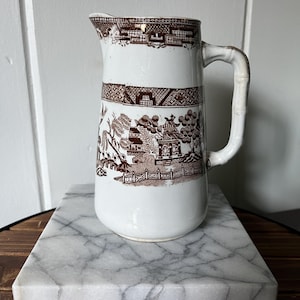 May include: A white ceramic pitcher with a brown transferware design featuring a landscape scene. The pitcher has a bamboo-style handle and is displayed on a marble surface. The pitcher is a classic piece of kitchenware.