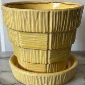 May include: A yellow ceramic planter with a matching saucer. The planter has a textured, stacked design, with a light brown rim. The saucer is round and has a similar textured pattern. The planter is suitable for indoor or outdoor use.