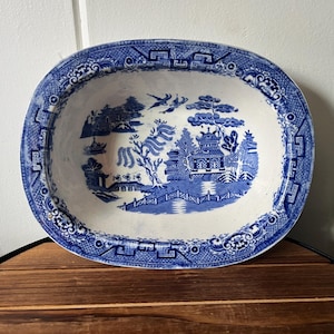 May include: An oval ceramic bowl featuring a blue and white Willow pattern. The design depicts a landscape with a pagoda, a bridge, a willow tree, and birds in flight. The rim is decorated with a geometric border pattern on a white background.