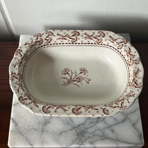 Può includere: Un piatto da portata ovale in ceramica bianco sporco con un motivo floreale marrone sul bordo e un motivo floreale al centro. Il piatto è posto su una superficie di marmo.