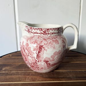 May include: A cream-coloured ceramic jug featuring a red transferware pattern depicting a rural landscape with trees, a horse-drawn cart, and a person. The rim is decorated with a continuous vine motif, and the handle is curved and solid.