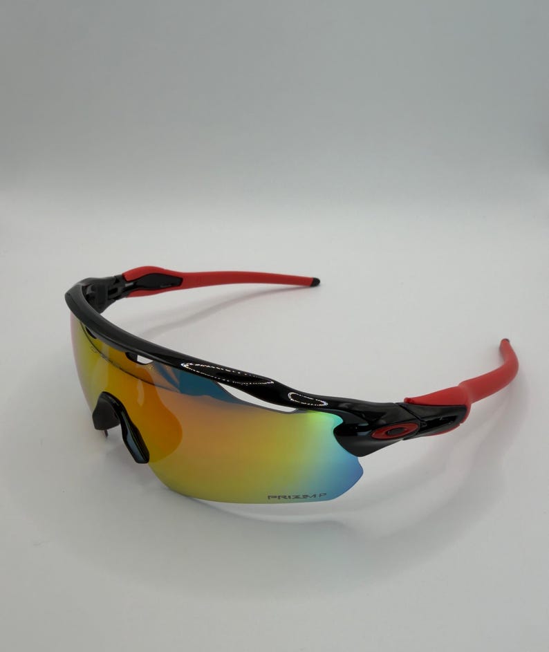 May include: Sport sunglasses with a black frame and red arms. The lenses have a gradient of colors, including yellow, orange, and blue. The brand name is visible on the lens.