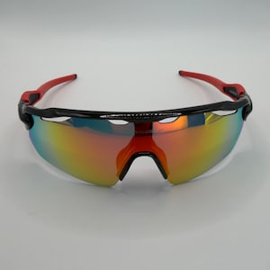 May include: Sport sunglasses with a black frame and red arms. The lenses have a mirrored finish with a gradient of colors, including yellow, orange, and blue. The sunglasses are designed for athletic activities.