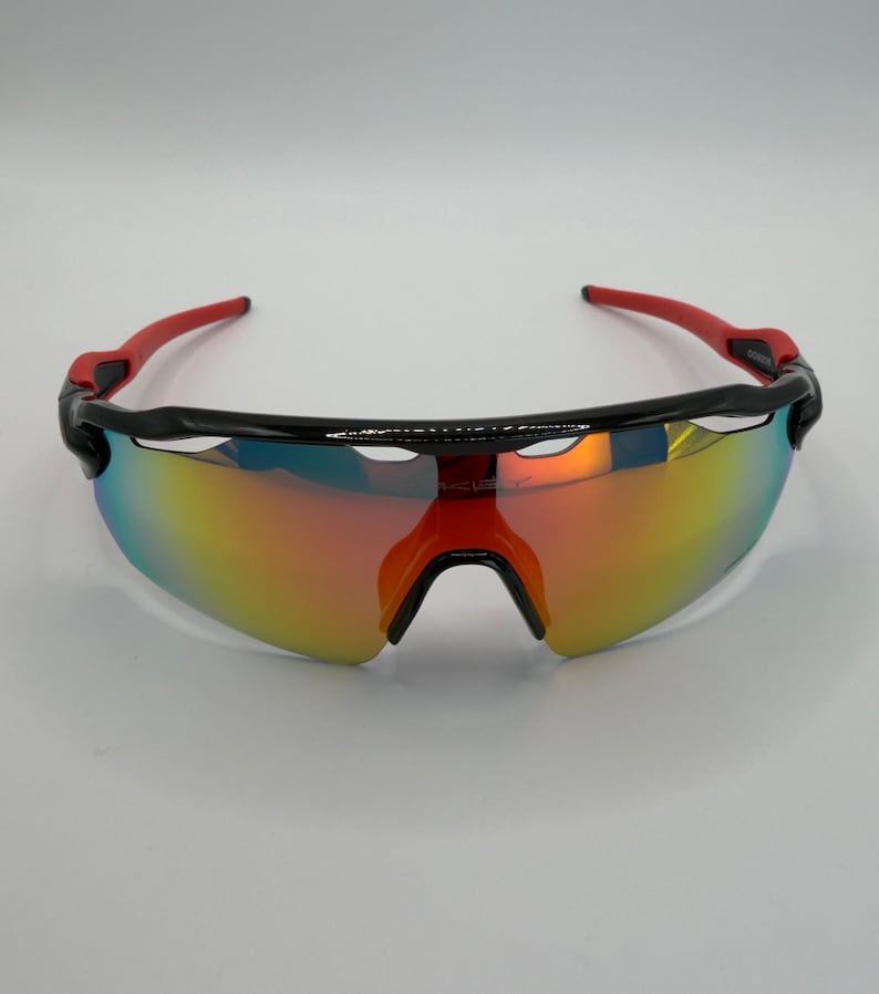 May include: Sport sunglasses with a black frame and red arms. The lenses have a mirrored finish with a gradient of colors, including yellow, orange, and blue. The sunglasses are designed for athletic activities.