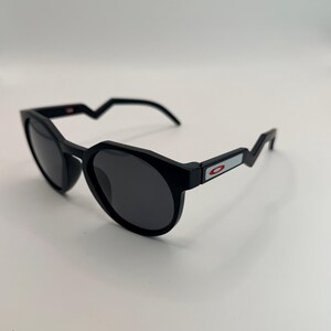 May include: Black sunglasses with a unique angular design. The frames are matte black with dark lenses. The temples have a zig-zag shape and a small rectangular accent with a red and white logo.