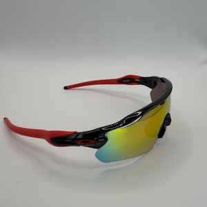 May include: Sport sunglasses with a black frame and red arms. The lenses have a yellow and blue iridescent finish. The sunglasses are designed for sports and outdoor activities.