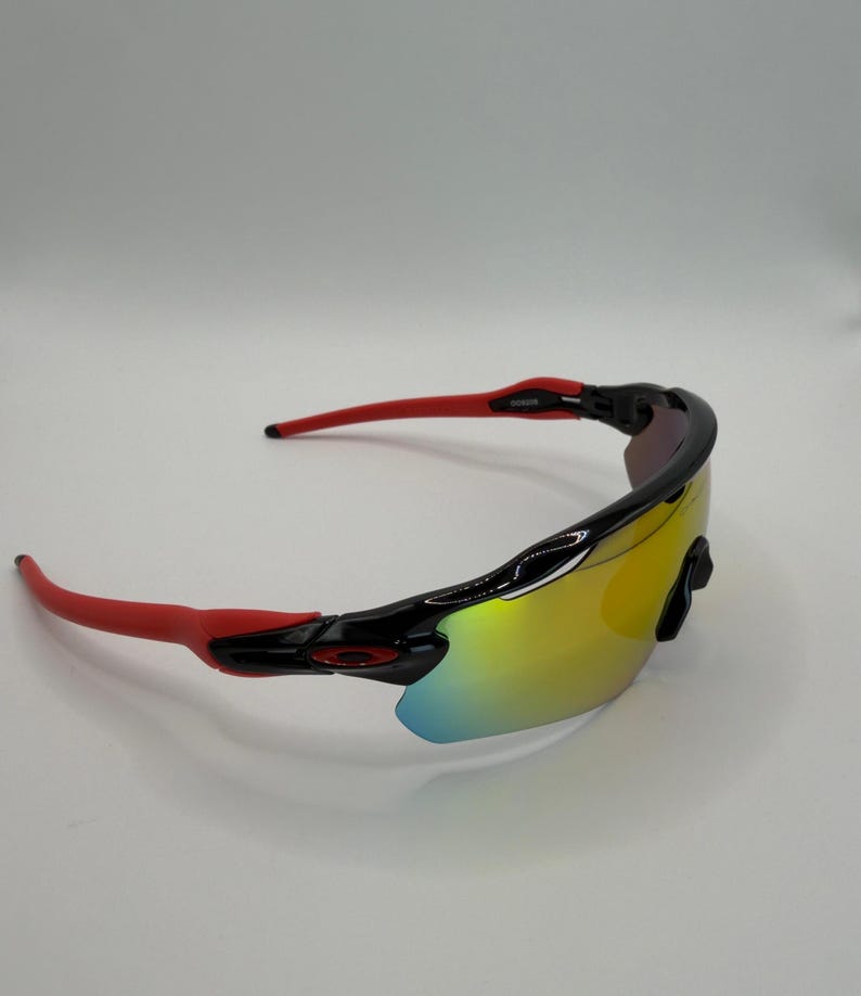 May include: Sport sunglasses with a black frame and red arms. The lenses have a yellow and blue iridescent finish. The sunglasses are designed for sports and outdoor activities.