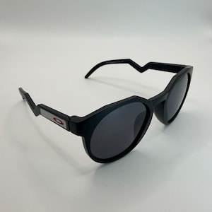 May include: Black sunglasses with a rounded frame and dark lenses. The temples have a unique angular design, and the Oakley logo is visible on the side. The sunglasses are set against a white background.