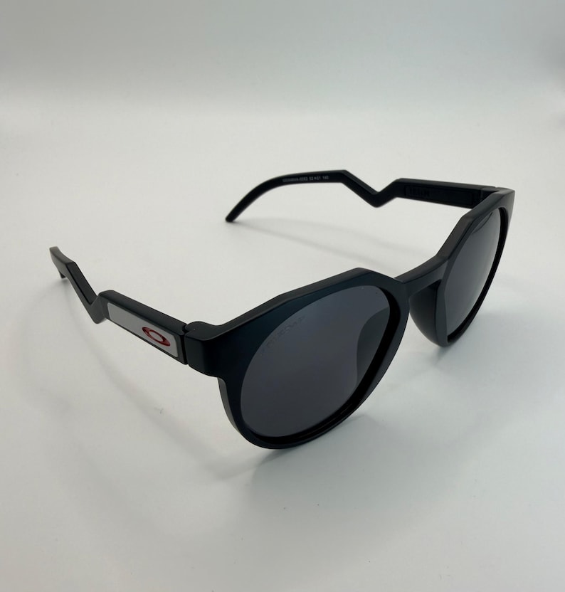 May include: Black sunglasses with a rounded frame and dark lenses. The temples have a unique angular design, and the Oakley logo is visible on the side. The sunglasses are set against a white background.