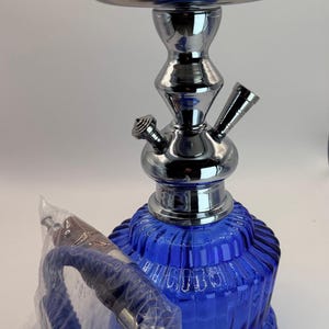 May include: A blue glass hookah with a chrome metal stem and bowl. The base has a textured design. A blue hose is in a plastic bag. Accessories are in the bowl.