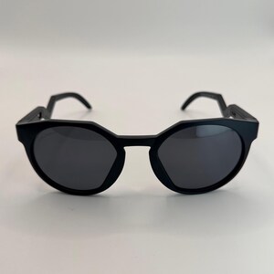 May include: Black sunglasses with a unique geometric frame design. The lenses are dark, and the arms have an angular shape. The sunglasses are set against a plain white background, highlighting their modern style.