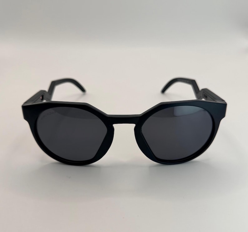 May include: Black sunglasses with a unique geometric frame design. The lenses are dark, and the arms have an angular shape. The sunglasses are set against a plain white background, highlighting their modern style.