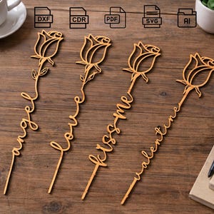 Könnte beinhalten: Vier Holzaufsätze in Rosenform mit Stielen. Jeder Aufsatz hat ein anderes Wort: "angel", "love", "kiss me" und "valentine". Die Aufsätze sind hellbraun und wahrscheinlich für Kuchendekorationen oder Bastelarbeiten gedacht.