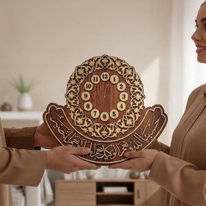 Könnte beinhalten: Eine Holz-Uhr mit arabischer Kalligraphie und einem dekorativen kreisförmigen Design. Das Zifferblatt hat die Zahlen 1 bis 12. Die Uhr ist braun mit goldenen Akzenten und wird von zwei Personen gehalten.