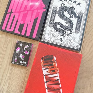 Może przedstawiać: Cztery albumy Stray Kids są wyświetlane na drewnianej powierzchni. Albumy są w różnych kolorach, w tym czarnym, czerwonym i białym. Okładki albumów zawierają grafiki i tekst, w tym słowa "MAX IDENTIFY" i "BINARY".