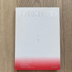 Op de afbeelding: Een wit boek met de titel "DARK BLOOD" in zilveren letters. De onderste rand loopt over van wit naar rood. Tekst en een puntkomma-symbool zijn ook zichtbaar. Het woord "ENHYPEN" staat rechtsonder gedrukt.