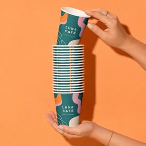 May include: A stack of teal paper cups with white, orange, and pink abstract shapes. The text LUNA CAFÉ COFFEE & GOOD VIBES is printed in white on the side of the cups. The cups are held vertically between two hands against a solid orange background.