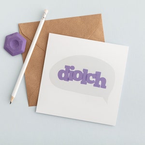 May include: A white greeting card with the word "diolch" in purple, inside a speech bubble. A brown envelope, a white pencil, and a purple eraser are also present, all on a light blue surface.