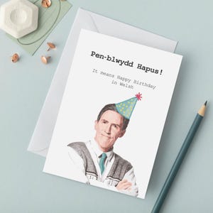 Puede incluir: Tarjeta de felicitación blanca con el texto galés « Pen-blwydd Hapus ! » y su traducción al inglés. La tarjeta presenta una ilustración de un hombre con un gorro de fiesta. Un lápiz turquesa está al lado.