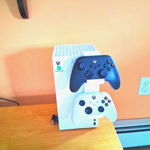 May include: A white Xbox Series S console with a controller stand holding a black and a white controller. The controllers have colored buttons and joysticks. The console is on a wooden surface against an orange wall.