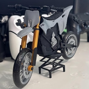 Op de afbeelding: Een miniatuurmotorfietsmodel in grijs en zwart, met goudkleurige voorvorken. Het model wordt gepresenteerd op een zwarte standaard, met details van de wielen, het frame en het stuur. De motorfiets is een gedetailleerde replica.