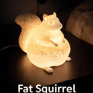 Könnte beinhalten: Eine Keramiklampe in Form eines pummeligen Eichhörnchens, von innen beleuchtet. Die Lampe hat einen flauschigen Schwanz und steht auf einer dunklen Oberfläche. Der Text "Fat Squirrel Lamp" ist darunter zu sehen.