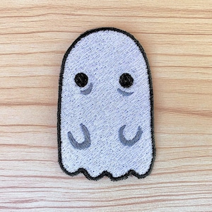 May include: A white ghost with black eyes and a small smile embroidered on a light brown background.