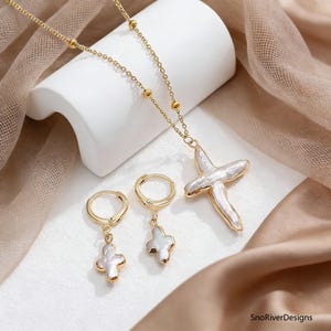 May include: A gold-coloured jewellery set featuring a necklace with a cross pendant and two pairs of hoop earrings. The cross and earring charms have a white, textured surface. The jewellery is displayed on a white surface and a draped, tan fabric.