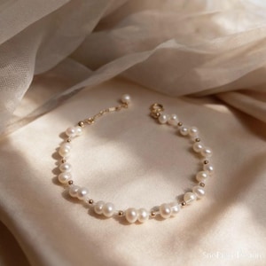 Könnte beinhalten: Ein zartes Armband mit weißen Perlen und kleinen goldenen Perlen. Das Armband ist an einer feinen Kette aufgereiht und wird auf einer cremefarbenen Satin-Oberfläche präsentiert. Der Schmuck ist für den täglichen Gebrauch konzipiert.