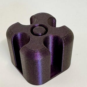 May include: A dark purple, 3D-printed object with a unique, geometric design. The object has a cylindrical base with four curved, vertical slots and a central circular indentation on top. The surface has a textured appearance.