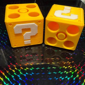 以下が含まれることがあります： 黄色い立方体のブロックが2つ。白いアウトラインのクエスチョンマークが描かれています。ブロックには円形と四角形の切り抜きがあります。ブロックは反射性のある虹色の表面に置かれています。