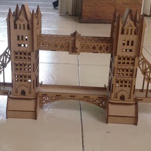May include: A wooden model of the Tower Bridge, featuring two towers connected by a central roadway and suspension bridges. The model is made of light brown wood and sits on a tiled surface.