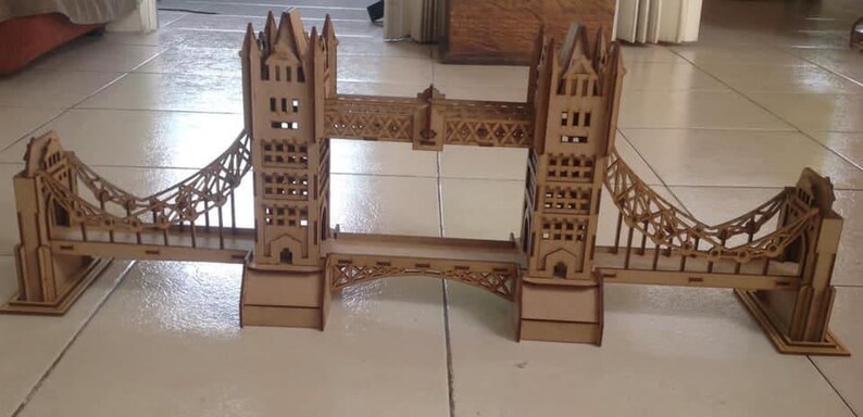 May include: A wooden model of the Tower Bridge, featuring two towers connected by a central roadway and suspension bridges. The model is made of light brown wood and sits on a tiled surface.