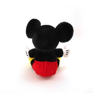 K&ouml;nnte beinhalten: Ein schwarzes und rotes Pl&uuml;sch-Mickey-Mouse-Spielzeug, das auf einer wei&szlig;en Oberfl&auml;che sitzt. Das Spielzeug hat gro&szlig;e schwarze Ohren und eine rote und gelbe Hose.