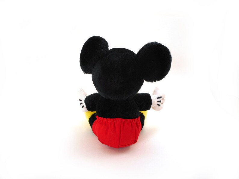 K&ouml;nnte beinhalten: Ein schwarzes und rotes Pl&uuml;sch-Mickey-Mouse-Spielzeug, das auf einer wei&szlig;en Oberfl&auml;che sitzt. Das Spielzeug hat gro&szlig;e schwarze Ohren und eine rote und gelbe Hose.