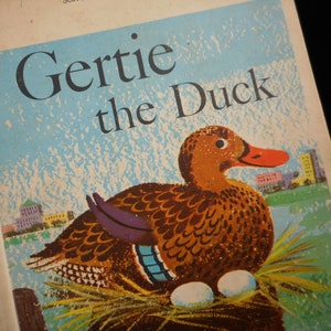 May include: Vintage children's book cover featuring a brown duck with a red beak and purple wing feathers. The title "Gertie the Duck" is in large blue letters against a light blue sky background with a city skyline.