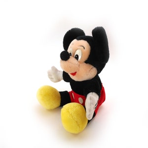 K&ouml;nnte beinhalten: Ein Pl&uuml;sch-Mickey-Mouse-Spielzeug mit schwarzem Fell, roten Shorts mit wei&szlig;en Punkten und gelben Schuhen. Das Spielzeug sitzt auf einer wei&szlig;en Oberfl&auml;che.