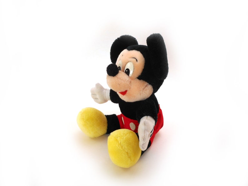 K&ouml;nnte beinhalten: Ein Pl&uuml;sch-Mickey-Mouse-Spielzeug mit schwarzem Fell, roten Shorts mit wei&szlig;en Punkten und gelben Schuhen. Das Spielzeug sitzt auf einer wei&szlig;en Oberfl&auml;che.